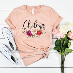 May include: A peach-colored t-shirt with the word "Chilena" printed in black script. The shirt features a floral design with red, pink, and white flowers.