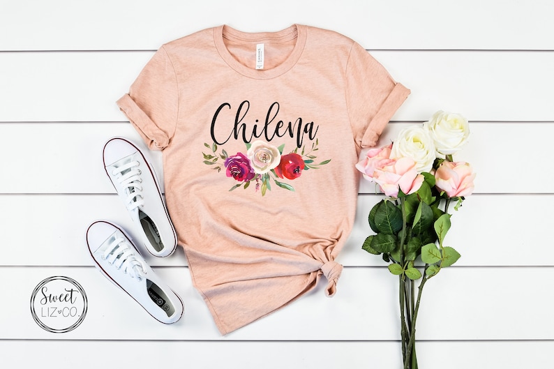 May include: A peach-colored t-shirt with the word "Chilena" printed in black script. The shirt features a floral design with red, pink, and white flowers.