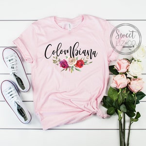 May include: Pink t-shirt with the text "Columbiana" in black script with a floral design underneath. The shirt is laid flat on a white wooden surface with a pair of white sneakers and a bouquet of pink roses.