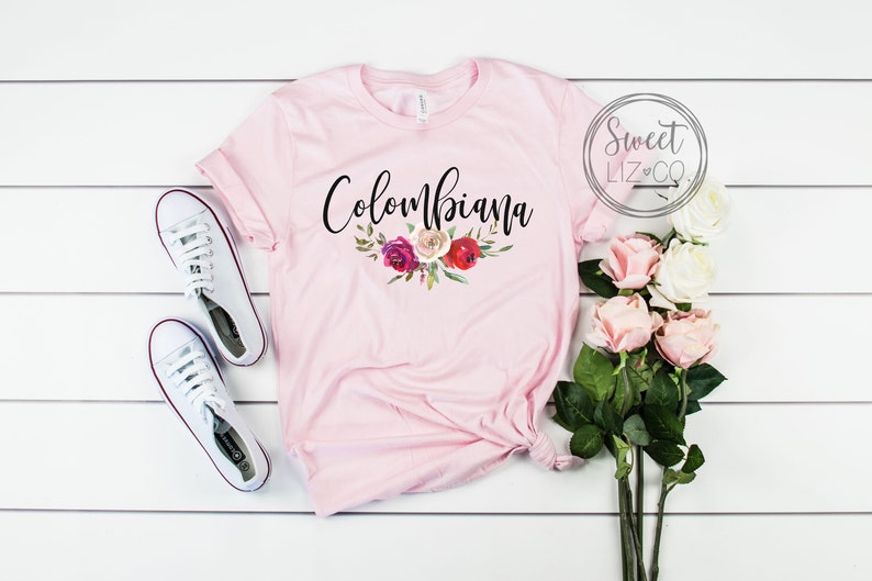 May include: Pink t-shirt with the text "Columbiana" in black script with a floral design underneath. The shirt is laid flat on a white wooden surface with a pair of white sneakers and a bouquet of pink roses.