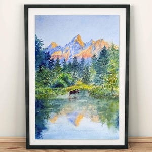 May include: Watercolour painting of a mountain landscape with a moose in a lake. The artwork features a black frame, a serene lake reflecting the mountains and trees, and a moose wading in the water. The mountains are illuminated with golden light.