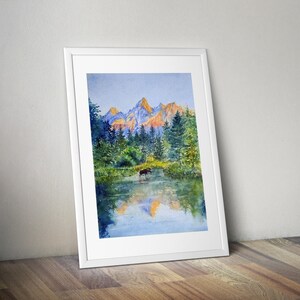 May include: Watercolour painting of a mountain landscape with a moose in a lake. The artwork features a vibrant colour palette of blues, greens, and oranges, framed in a white border. The painting is leaning against a wooden floor.