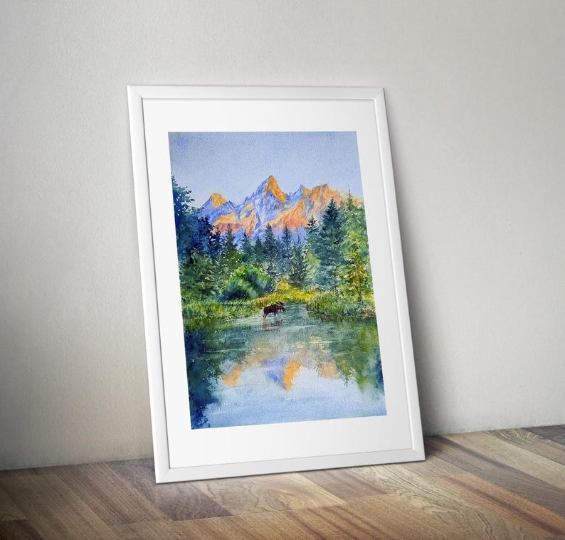 May include: Watercolour painting of a mountain landscape with a moose in a lake. The artwork features a vibrant colour palette of blues, greens, and oranges, framed in a white border. The painting is leaning against a wooden floor.