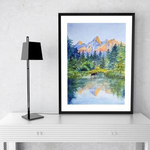 May include: Watercolour painting of a mountain landscape with a moose in a lake. The artwork features a black frame and is displayed on a white table. A black lamp stands to the left of the painting.