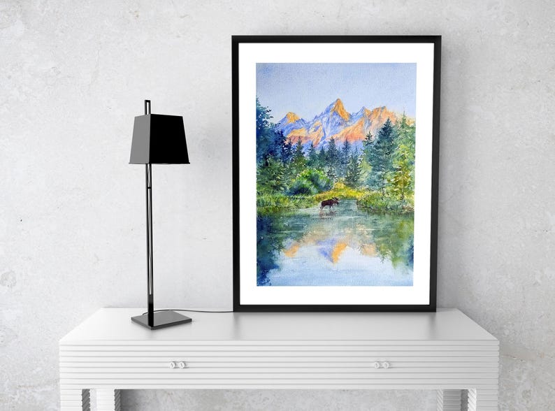 May include: Watercolour painting of a mountain landscape with a moose in a lake. The artwork features a black frame and is displayed on a white table. A black lamp stands to the left of the painting.