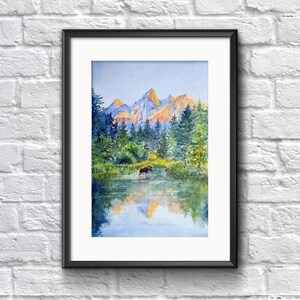 May include: Watercolour painting of a mountain landscape with a lake and trees. The mountains are in the background with orange and yellow hues. A moose stands in the water. The painting is framed in a black frame.