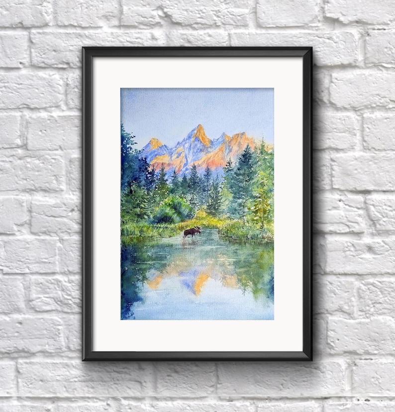 May include: Watercolour painting of a mountain landscape with a lake and trees. The mountains are in the background with orange and yellow hues. A moose stands in the water. The painting is framed in a black frame.