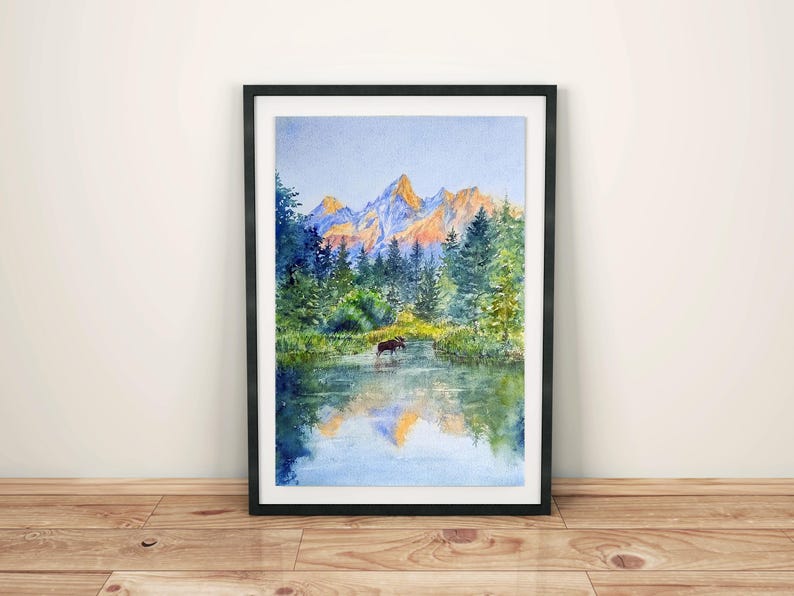May include: Watercolour painting of a mountain landscape with a moose in a lake. The artwork features a black frame, a serene lake reflecting the mountains and trees, and a moose wading in the water. The mountains are illuminated with golden light.
