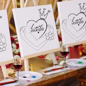 May include: Three white canvases on easels with the phrase "Love Yourself" written in black lettering on each canvas. The canvases are set up on a wooden table with wine glasses and paint supplies.