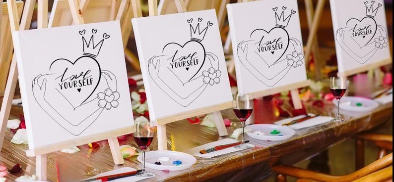 May include: Three white canvases on easels with the phrase "Love Yourself" written in black lettering on each canvas. The canvases are set up on a wooden table with wine glasses and paint supplies.