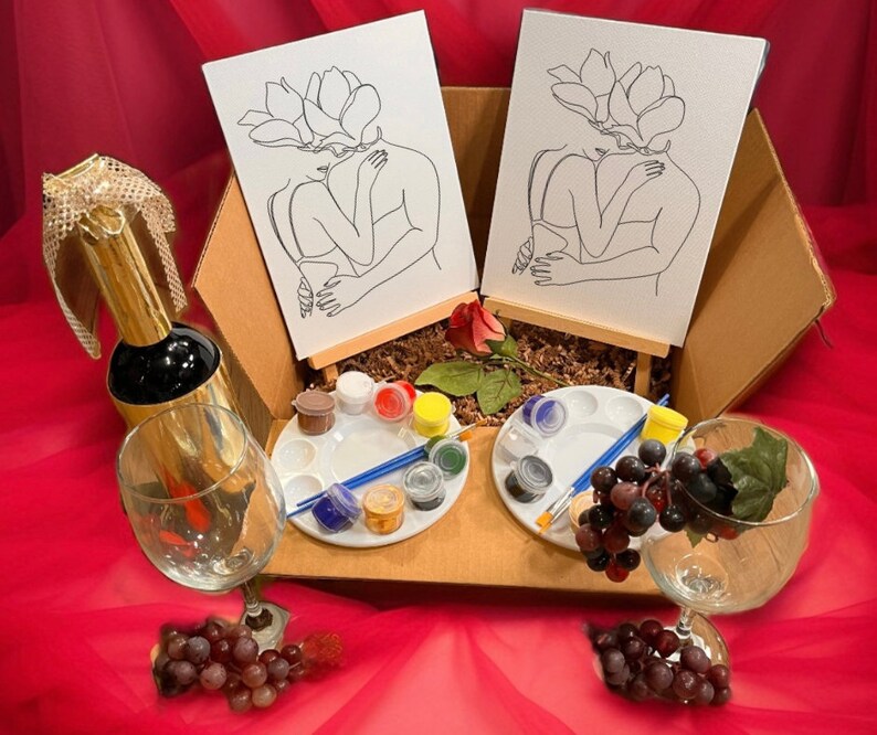 May include: A gift set in a cardboard box, featuring two canvases with line art of embracing figures, paint palettes, brushes, and a bottle of champagne. Also includes two wine glasses and grapes, all on a red background.