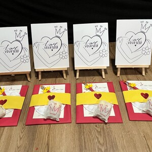 May include: Four white canvases on easels with the phrase "Love Yourself" and a crown above a heart. Each canvas is accompanied by a red and yellow gift bag with a paint palette, paintbrushes, and a small bag with a crown.