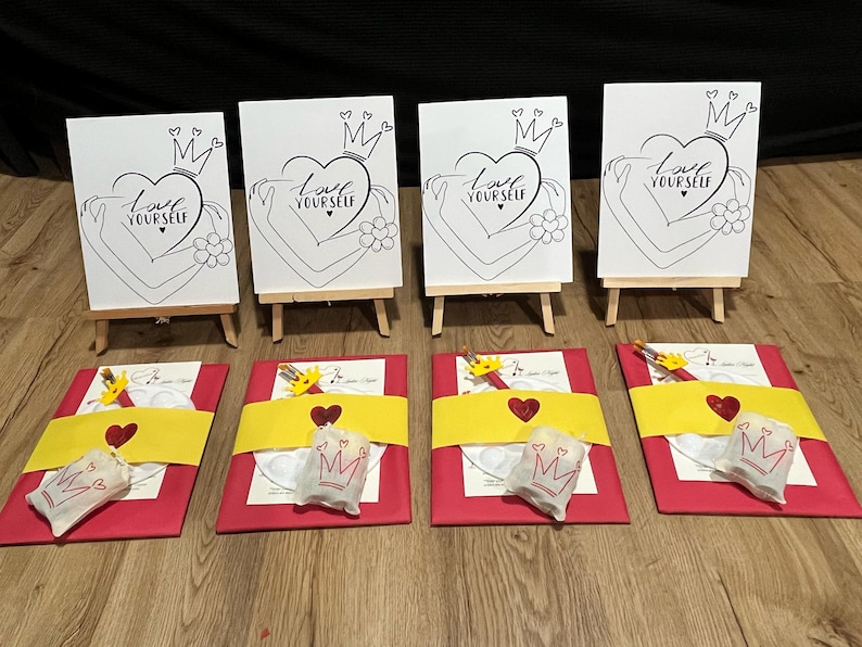 May include: Four white canvases on easels with the phrase "Love Yourself" and a crown above a heart. Each canvas is accompanied by a red and yellow gift bag with a paint palette, paintbrushes, and a small bag with a crown.