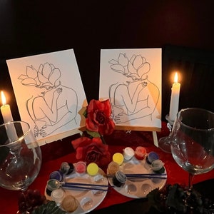May include: Two canvases with line art of embracing figures with flower-shaped heads, set on easels. The scene includes paint palettes, brushes, red roses, wine glasses, and lit candles, creating a romantic ambiance.