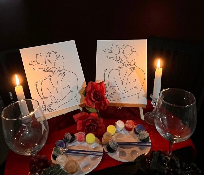 May include: Two canvases with line art of embracing figures with flower-shaped heads, set on easels. The scene includes paint palettes, brushes, red roses, wine glasses, and lit candles, creating a romantic ambiance.