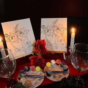 May include: Two canvases with line art of roses and figures, set on small easels. Paint palettes with various colors, paintbrushes, wine glasses, and lit candles create a romantic atmosphere. Red roses add to the decor.
