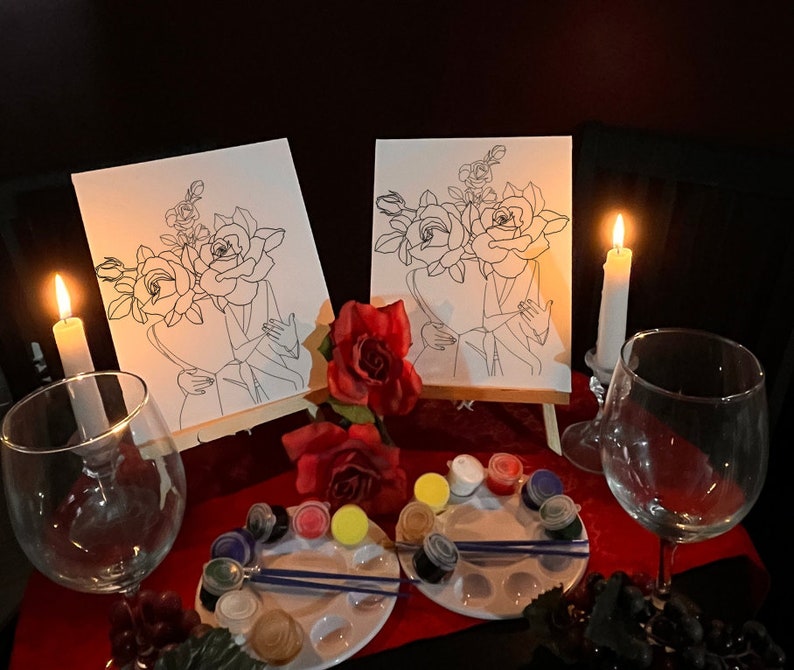 May include: Two canvases with line art of roses and figures, set on small easels. Paint palettes with various colors, paintbrushes, wine glasses, and lit candles create a romantic atmosphere. Red roses add to the decor.