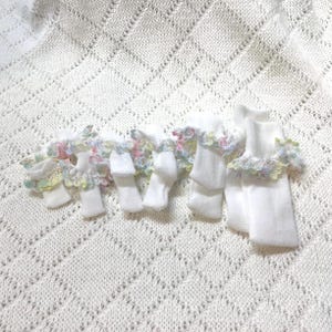 May include: A set of eight white baby socks with colorful floral lace trim. The socks are laid out on a white crocheted blanket.