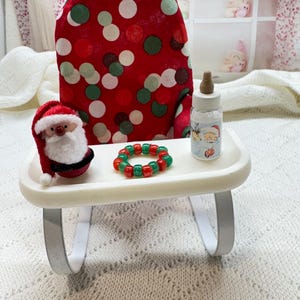 May include: A miniature high chair with a red fabric seat featuring white, red, and green polka dots. On the white tray sits a small Santa Claus figurine, a red and green beaded ring, and a miniature baby bottle with a Santa graphic.