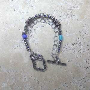 May include: A silver-toned bracelet with a toggle clasp. The bracelet features a chain design with various beads in shades of gray, blue, and maroon. The clasp is a decorative four-leaf clover shape.