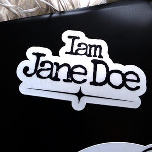 May include: A white die-cut sticker with a black border and black text that reads I am Jane Doe in a stylised, slightly distressed font. Below the text is a thin horizontal line featuring a centred four-pointed star shape.