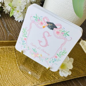 May include: A stack of square graduation announcement cards with a floral and ribbon design. The cards feature a pink letter "S" and a graduation cap. The cards are displayed on a gold tray with a clear stand.