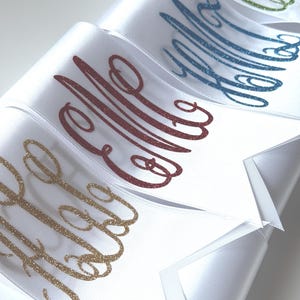 May include: A series of white satin ribbons arranged in a row, each featuring a unique monogram in cursive script. The monograms are applied in glittery finishes of gold, dark red, blue, and green, creating a textured, sparkling effect.