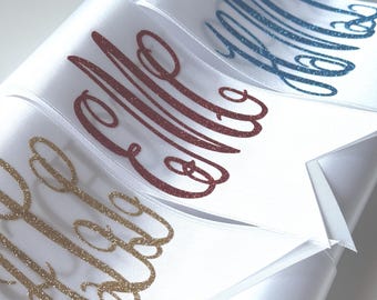 May include: A series of white satin ribbons featuring personalized monograms in glittery script lettering. The monograms appear in various colors including gold, dark red, blue, and green, each applied to the smooth surface of the fabric.