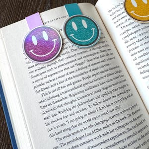 May include: Three smiley face bookmarks in purple, teal, and yellow, each with a white outline and a happy face design. The bookmarks are placed in an open book with text visible.