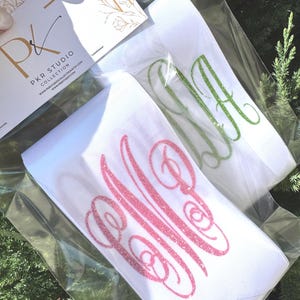 May include: Two white elastic headbands packaged in clear plastic, each featuring a glittery monogram. One headband displays a pink glitter monogram, while the other features a green glitter monogram. A PKR Studio Collection card is attached.