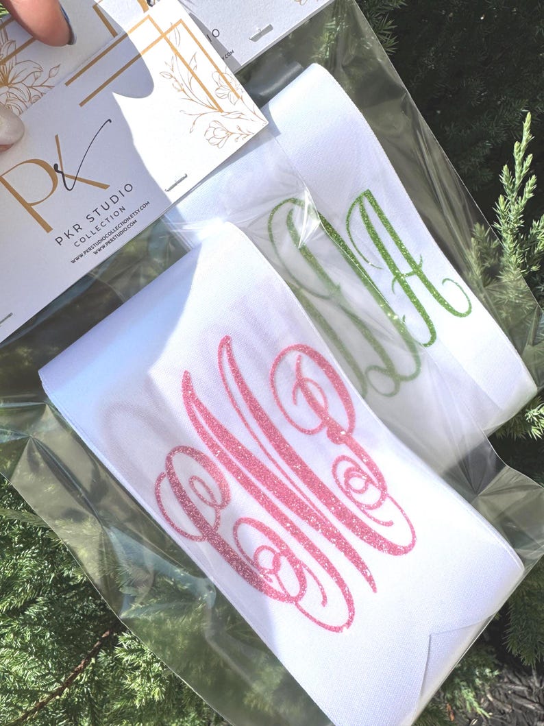May include: Two white elastic headbands packaged in clear plastic, each featuring a glittery monogram. One headband displays a pink glitter monogram, while the other features a green glitter monogram. A PKR Studio Collection card is attached.