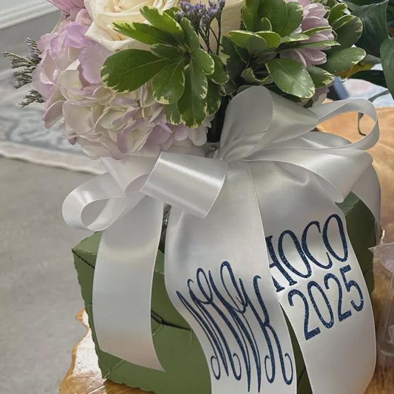 May include: A floral arrangement featuring light pink and white flowers with green leaves, set in a square olive green container. A wide white satin ribbon is tied around the container, printed with a dark blue monogram and the text HOCO 2025.