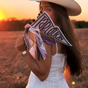 May include: A white cowboy hat and a triangular felt pennant flag featuring the text Tarleton in black lettering. The pennant is decorated with an assortment of hanging ribbons in shades of purple, black, and white, including lace and gingham patterns.