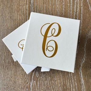 May include: Two square white gift boxes with a white ribbon pull tab. Each box features a large, metallic gold cursive letter C printed in the center of the lid. The boxes are stacked slightly offset, revealing the top and side surfaces.