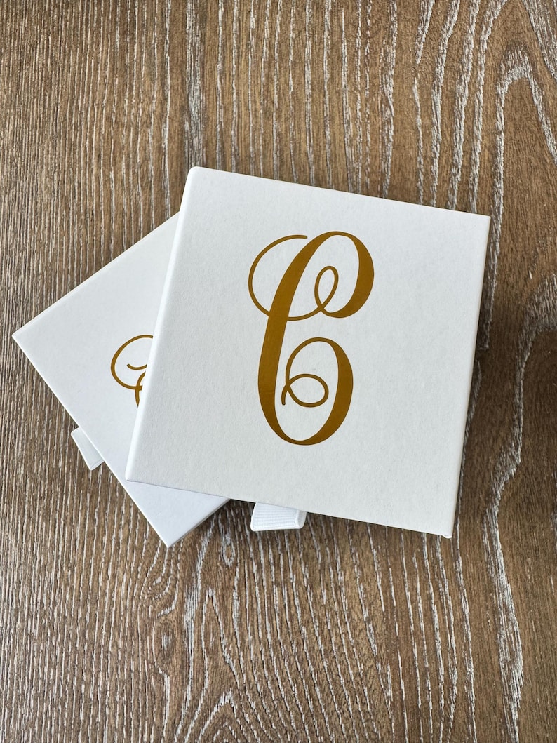 May include: Two square white gift boxes with a white ribbon pull tab. Each box features a large, metallic gold cursive letter C printed in the center of the lid. The boxes are stacked slightly offset, revealing the top and side surfaces.