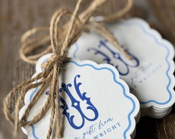 May include: A stack of scalloped white gift tags with a blue border and a blue decorative monogram. The text reads a gift from LORI WRIGHT. The tags are tied together with a brown jute twine bow, creating a rustic and functional presentation.