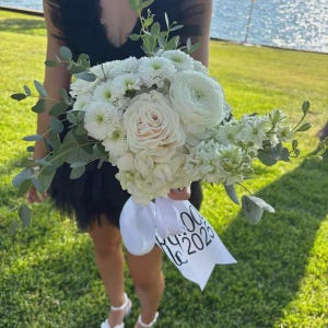 May include: A floral bouquet featuring white roses, ranunculus, and chrysanthemums accented with green eucalyptus leaves. A white ribbon is tied around the stems, printed with the black text "2025" and additional partially visible lettering.