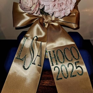 May include: A gold satin ribbon tied in a bow, featuring dark green glitter lettering. The left tail displays the monogram LSA, while the right tail features the text HOCO 2025. Pale pink artificial flowers are tucked behind the center knot.