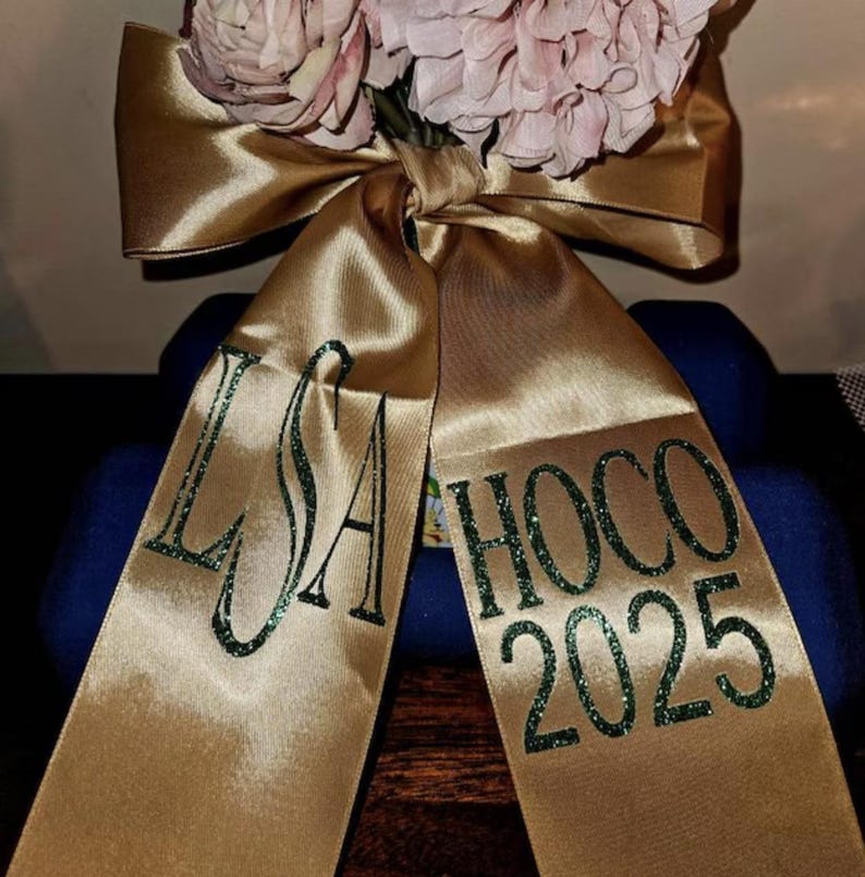 May include: A gold satin ribbon tied in a bow, featuring dark green glitter lettering. The left tail displays the monogram LSA, while the right tail features the text HOCO 2025. Pale pink artificial flowers are tucked behind the center knot.