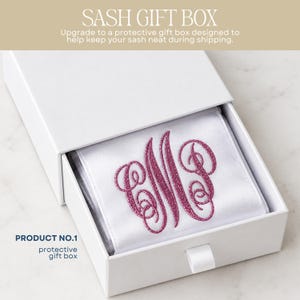 May include: A white rectangular sliding gift box containing a white satin sash with a large, glittery pink embroidered monogram featuring the letters M and P. The box lid displays the text SASH GIFT BOX and product information.