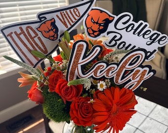 May include: A vibrant floral arrangement in a clear glass vase. The bouquet features red roses, orange gerbera daisies, and other colorful blooms. Decorative signs with the text "The Ville", "College Bound", and "Keely" are included.