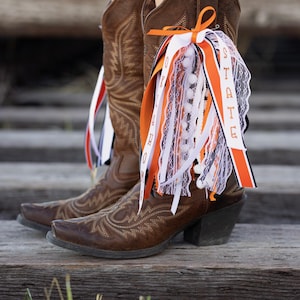 May include: Brown leather cowboy boots with decorative stitching and a pointed toe. The boots are adorned with orange, white, and black ribbons, some with the word "STATE" printed on them. The boots are on a wooden surface.