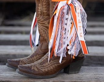 May include: Brown leather cowboy boots with decorative stitching and a pointed toe. The boots are adorned with orange, white, and black ribbons, some with the word "STATE" printed on them. The boots are on a wooden surface.