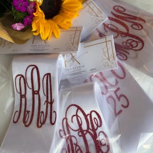 May include: White satin sashes featuring red glitter monogram lettering and the number 25, packaged in clear plastic sleeves with P&R Studio Collection branded cards, displayed alongside a bouquet of yellow sunflowers and purple flowers.