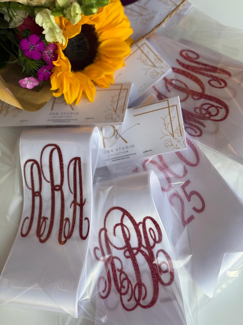 May include: White satin sashes featuring red glitter monogram lettering and the number 25, packaged in clear plastic sleeves with P&R Studio Collection branded cards, displayed alongside a bouquet of yellow sunflowers and purple flowers.