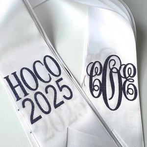 May include: A white satin sash featuring dark blue glitter text that reads HOCO 2025 on one side and a dark blue glitter monogram with three intertwined letters on the other side. The sash has a smooth, shiny finish and pointed ends.