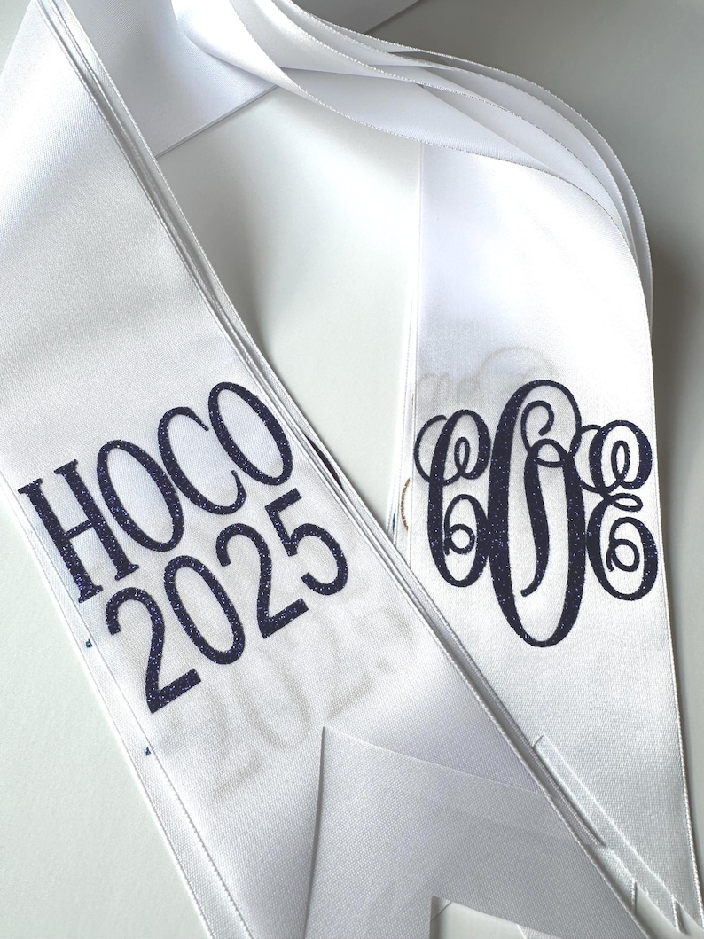May include: A white satin sash featuring dark blue glitter text that reads HOCO 2025 on one side and a dark blue glitter monogram with three intertwined letters on the other side. The sash has a smooth, shiny finish and pointed ends.