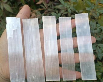 May include: Five white selenite sticks, also known as selenite wands, are arranged in a row on a hand. The sticks are smooth and have a natural, striated texture.
