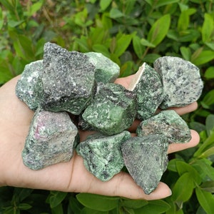 Op de afbeelding: Een verzameling ruwe groene en zwarte edelstenen, sommige met een vleugje roze, in een hand.