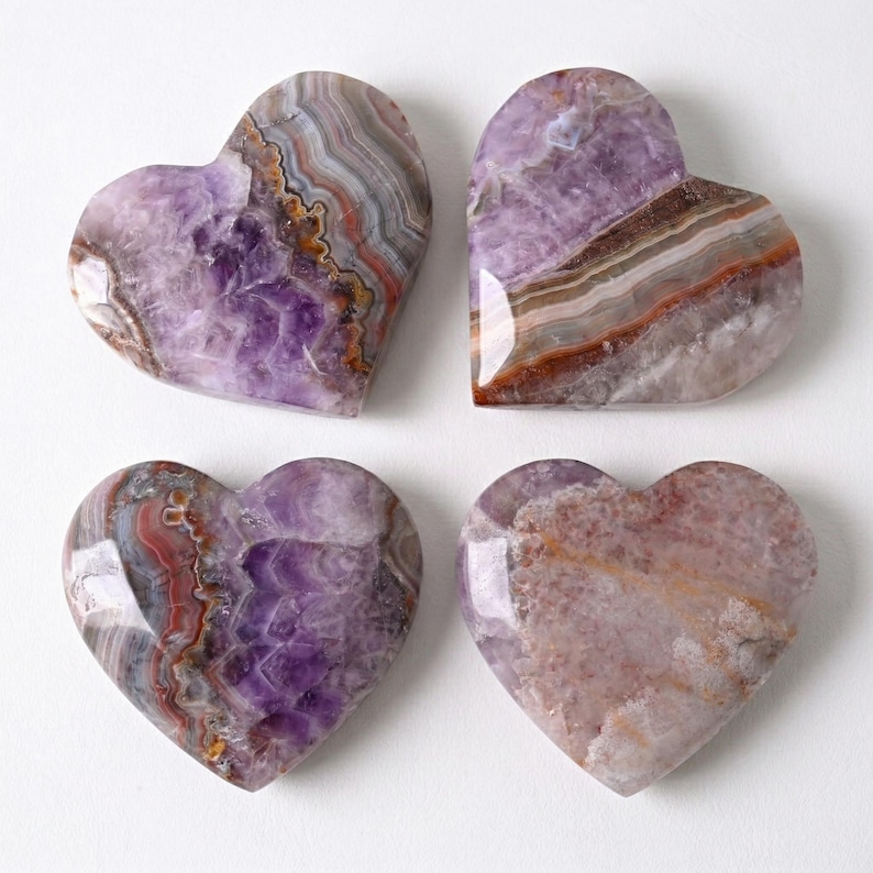May include: Four heart-shaped stones with polished surfaces. The stones display a range of colors, including shades of purple, brown, gray, and white, with visible banding and patterns. The arrangement is on a white background.